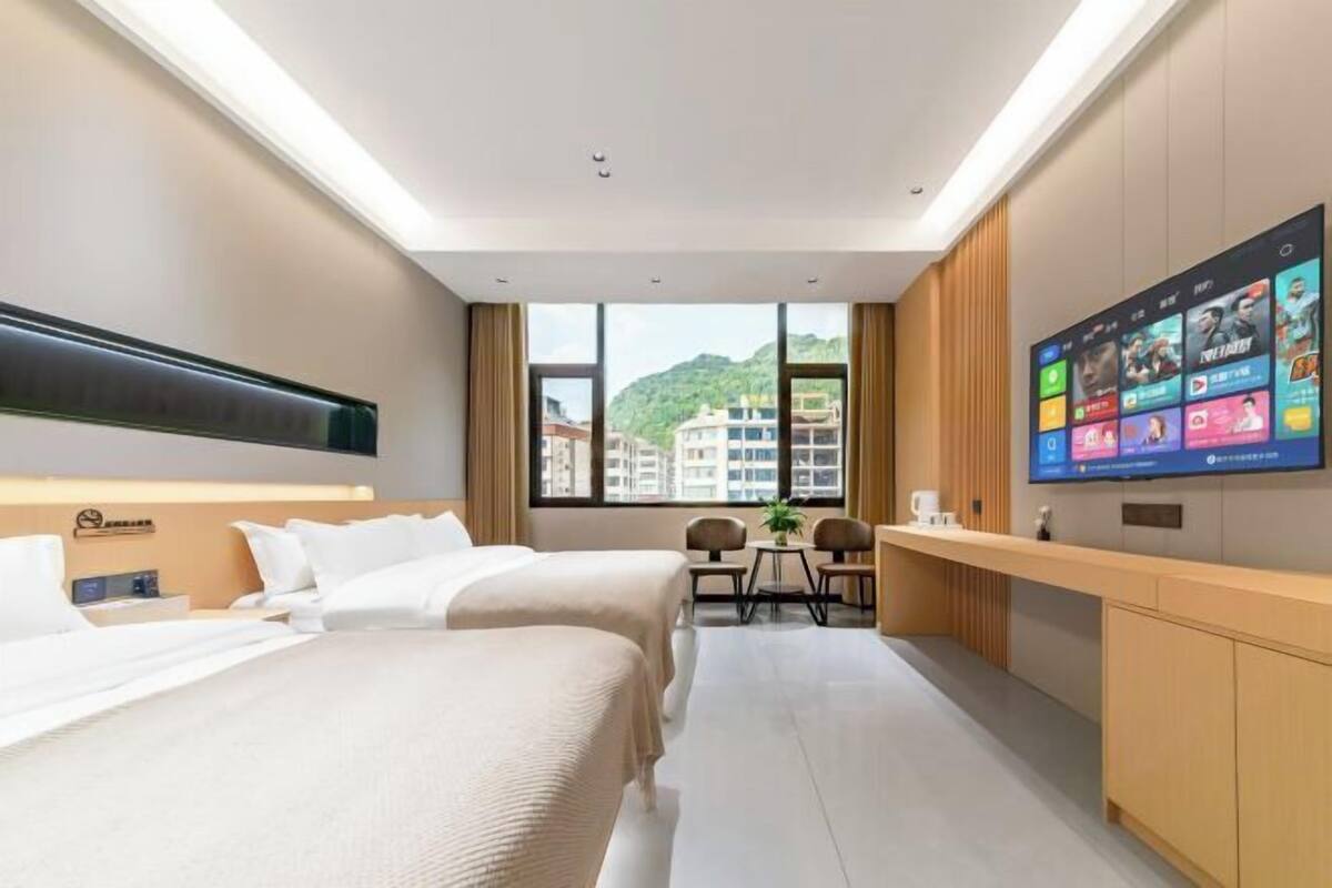A hotel room with two beds, a desk, a TV, and a window with a view of buildings and hills.