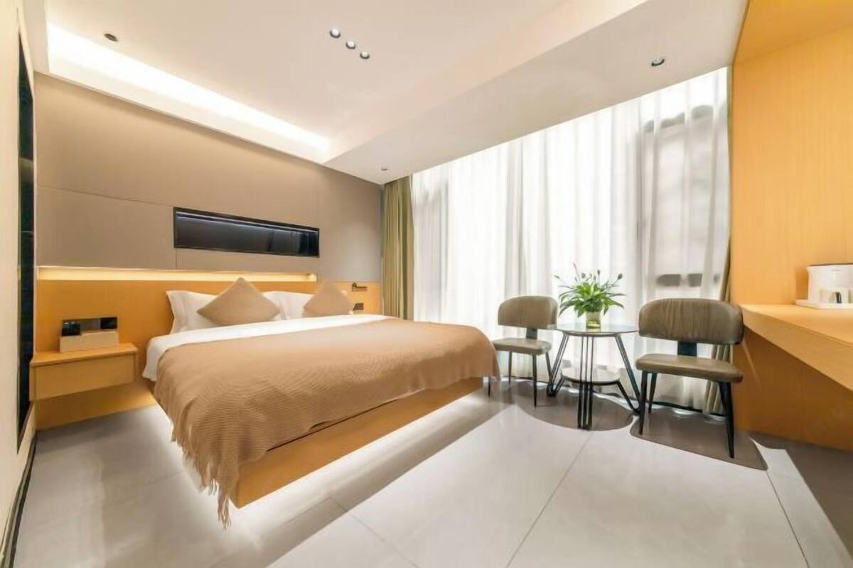 A modern hotel room with a large bed, bedside tables, a flat-screen TV, a small round table with two chairs, and a large window with curtains.