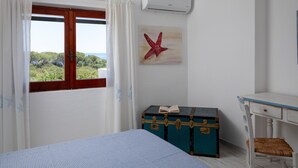 A bedroom with a bed, a window with curtains, a wall-mounted air conditioner, a wooden desk, a chair, and a blue trunk with a book on it. - Casa dell'Asfodelo by Wonderful Italy (Budoni)
