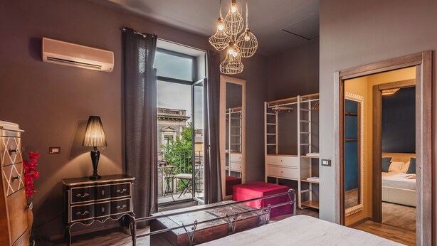 A modern bedroom with a large bed, a wardrobe, a vanity, and a window with a view of a building and greenery. - Ursino apartment with balcony by Wonderful Italy (Catania)