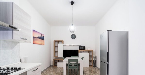 A compact kitchen with a gas stove, a refrigerator, a microwave, a dining table with chairs, a pendant light, and a cityscape painting on the wall.