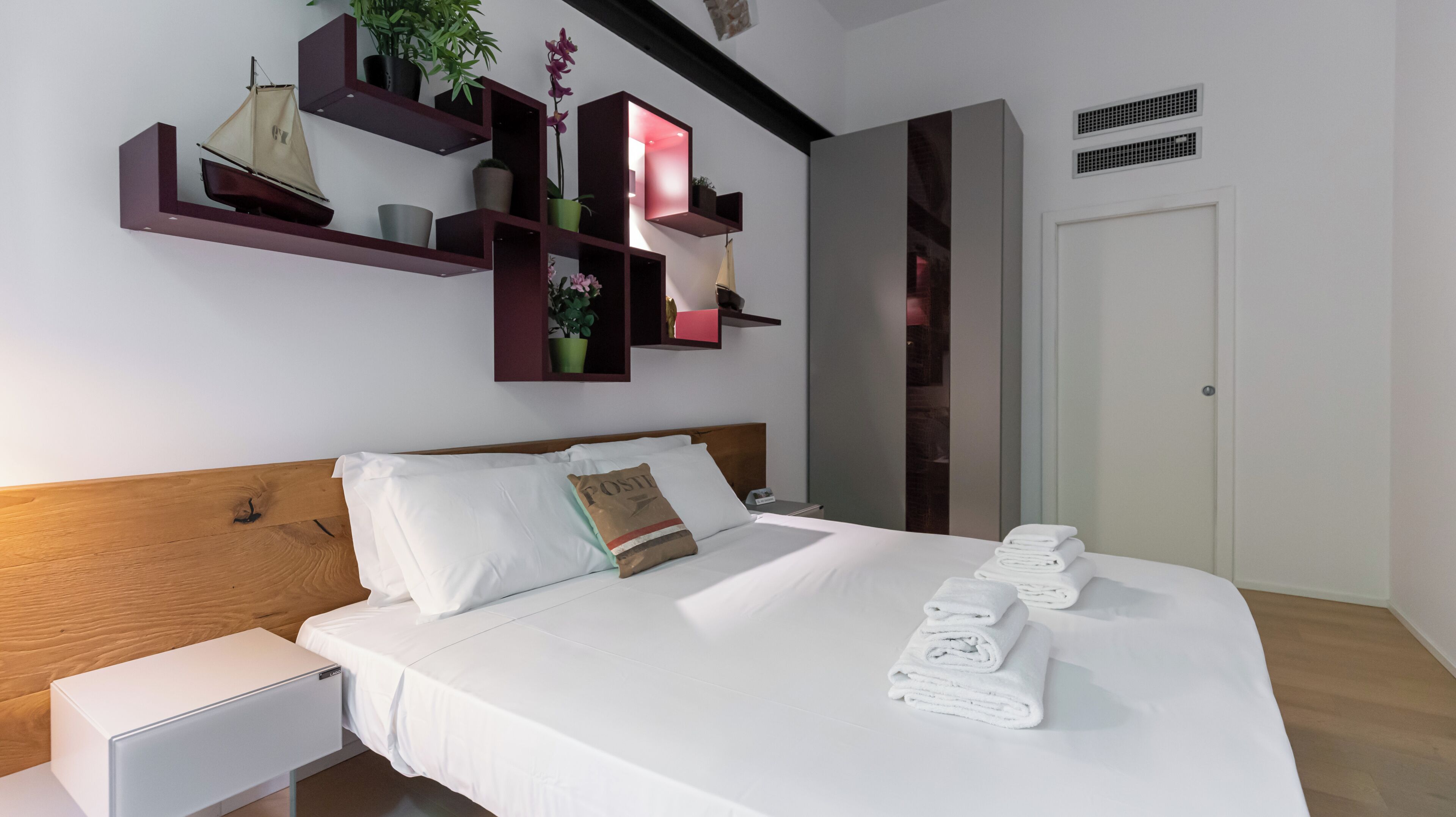 Habitación de hotel moderna con una cama bien hecha, una mesita de noche, un escritorio pequeño y un estante de pared con objetos decorativos.