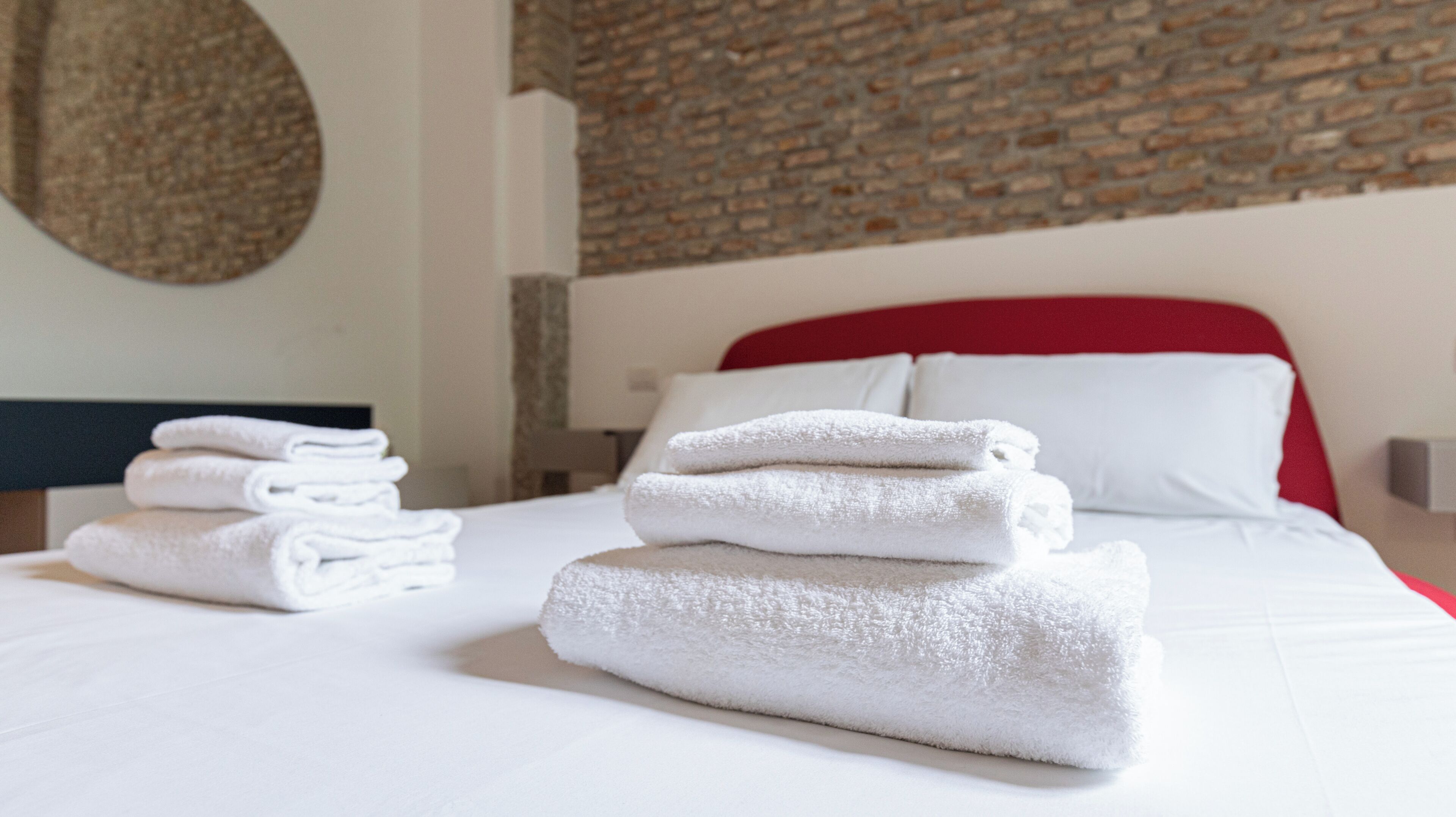 A neatly made bed with white linens and a red headboard, a stack of folded towels, and a brick wall in the background.
