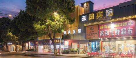 A street view of a commercial area with illuminated storefronts, including a restaurant and a shop with signage in a foreign language.