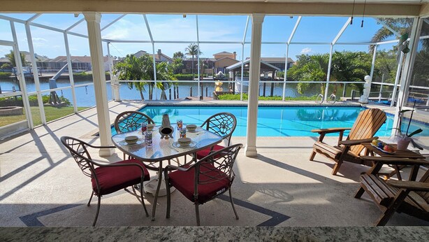 A covered outdoor area with a pool, a table with chairs, and a view of a body of water and buildings in the background. - Tropical Waterfront Pool Home, Private Dock, Kayaks, Direct Access to Tampa Bay! (Apollo Beach)