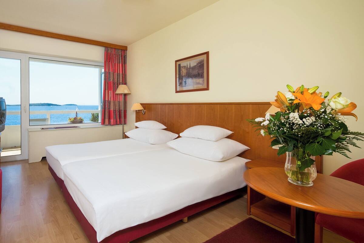 A hotel room with a bed, a desk with a telephone, a chair, a table with a vase of flowers, a window with a view of the sea, and a painting on the wall.