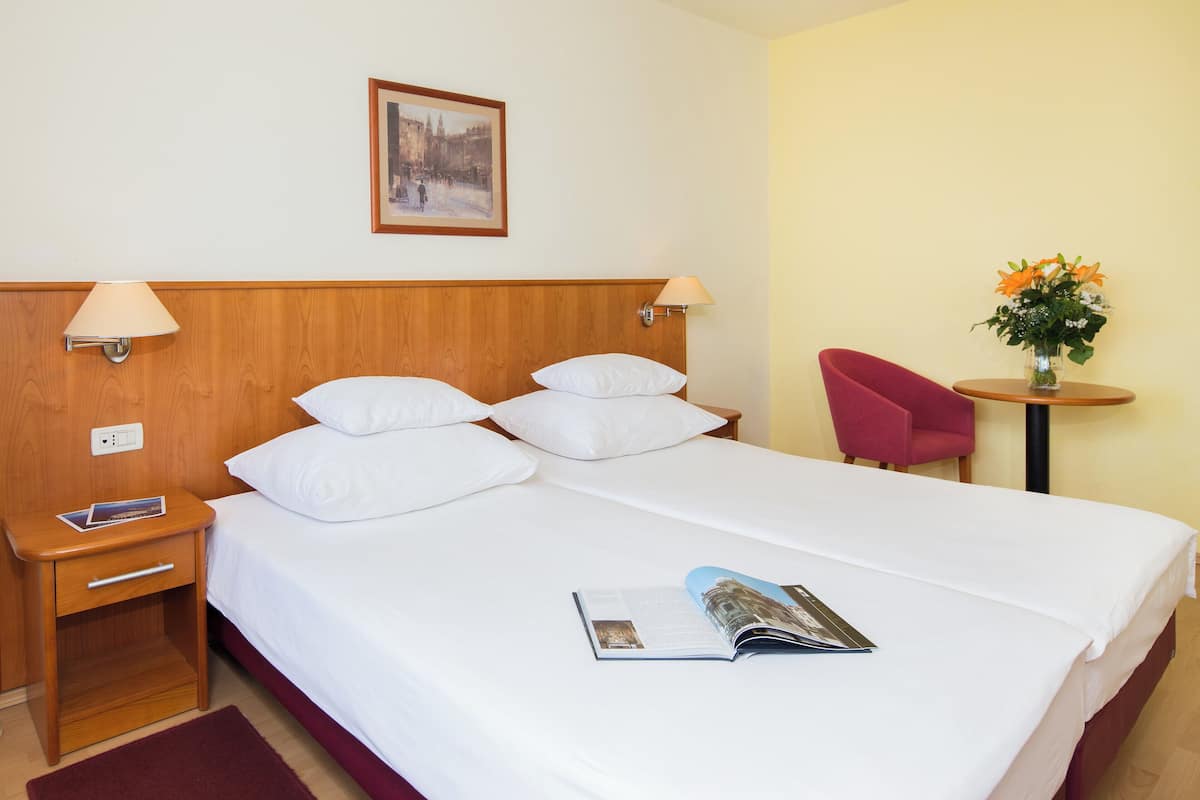 A hotel room with a bed, two bedside lamps, a nightstand, a magazine, a red chair, a small table with a vase of flowers, and a framed picture on the wall.