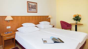 Una camera d'albergo con un letto, due abat-jour, un comodino, una rivista, una poltrona rossa, un tavolino con un vaso di fiori e un quadro appeso al muro.