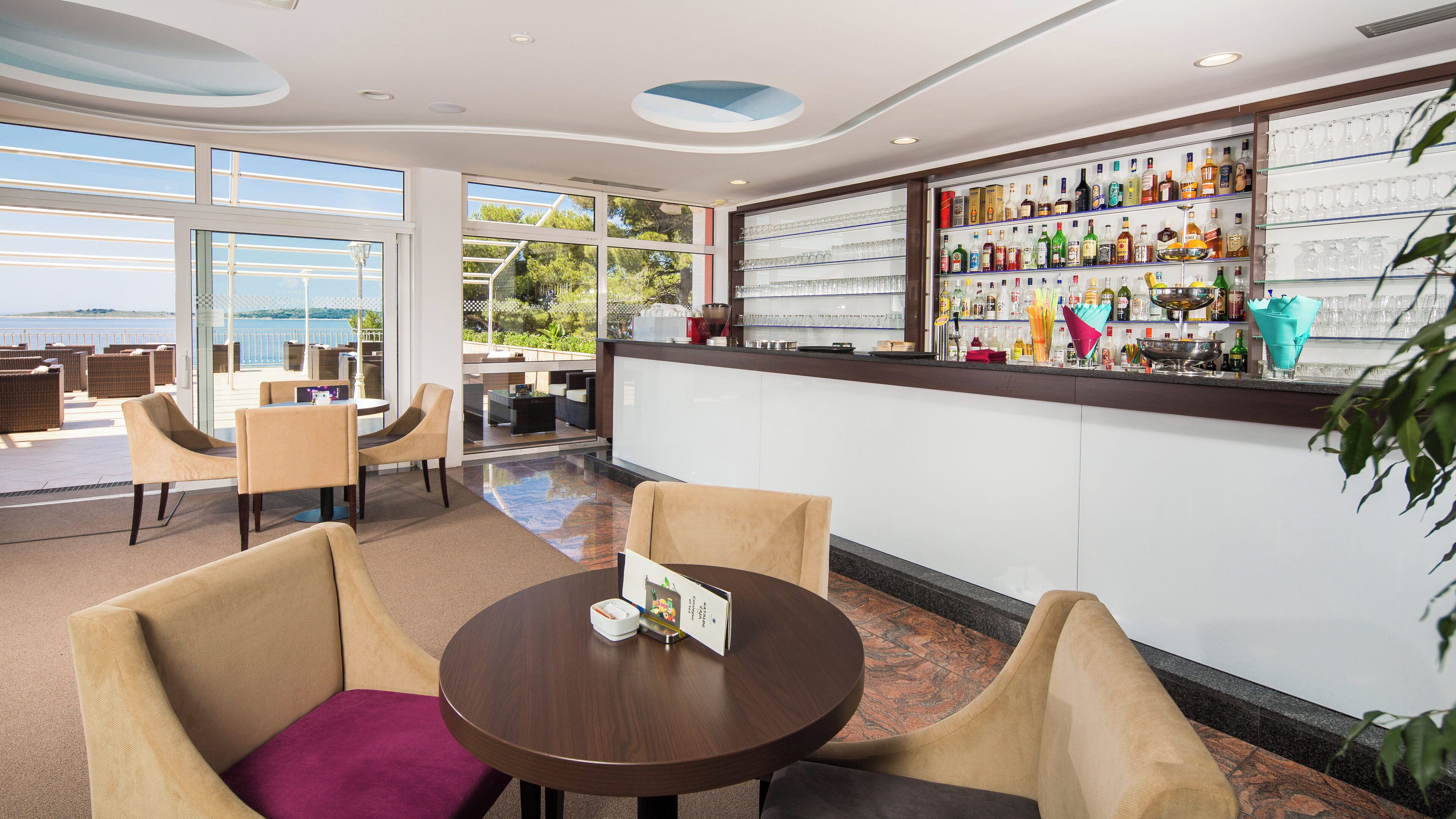A bar area with a view of the sea, seating with a purple cushion, and a variety of liquor bottles and glasses on shelves.