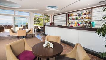 A bar area with a view of the sea, seating with a purple cushion, and a variety of liquor bottles and glasses on shelves.