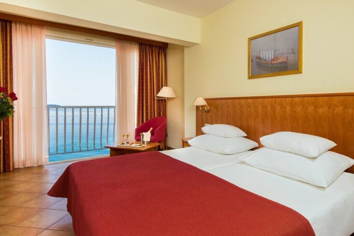 A hotel room with a large bed, a wooden headboard, a balcony with curtains, a red chair, and a painting on the wall.