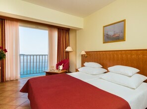 Habitación de hotel con una cama grande, un cabecero de madera, un balcón con cortinas, una silla roja y un cuadro en la pared.