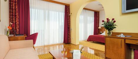 A hotel room with a sofa, a wooden desk, a TV, a bed, and a window with curtains.