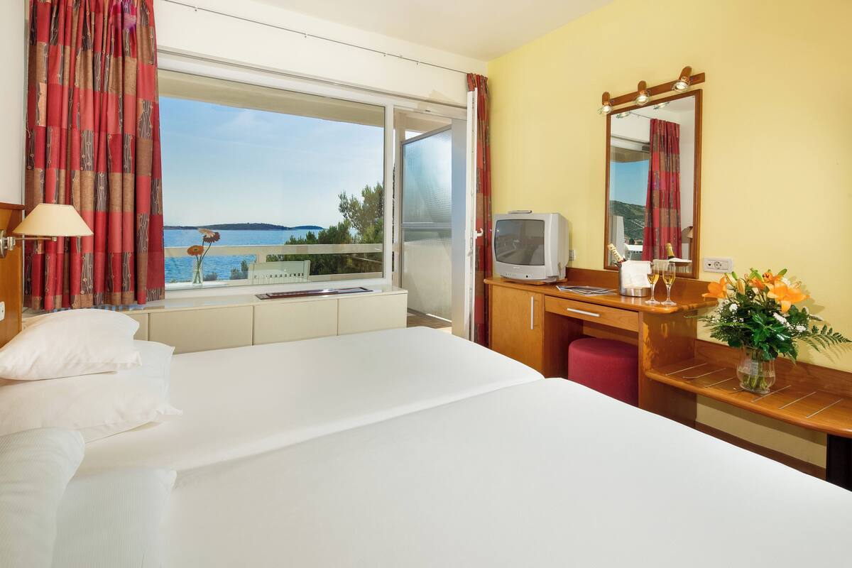 A hotel room with a bed, a desk, a television, a mirror, and a view of the sea.