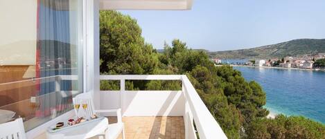 A balcony with white furniture, a table with a glass of drink, and a scenic view of a coastal town and water.