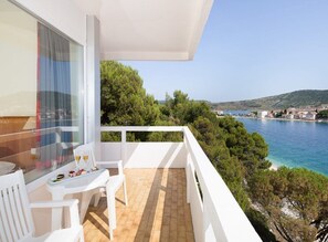 Un balcon agrémenté de meubles blancs, une table avec un verre, et une vue panoramique sur une ville côtière et la mer.