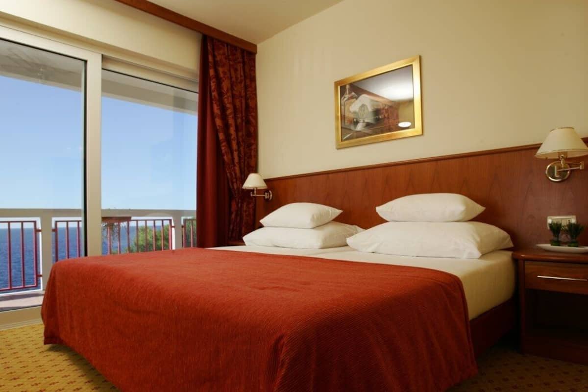 A hotel room with a large bed, a wooden headboard, a nightstand with a lamp, and a window with a view of the sea.