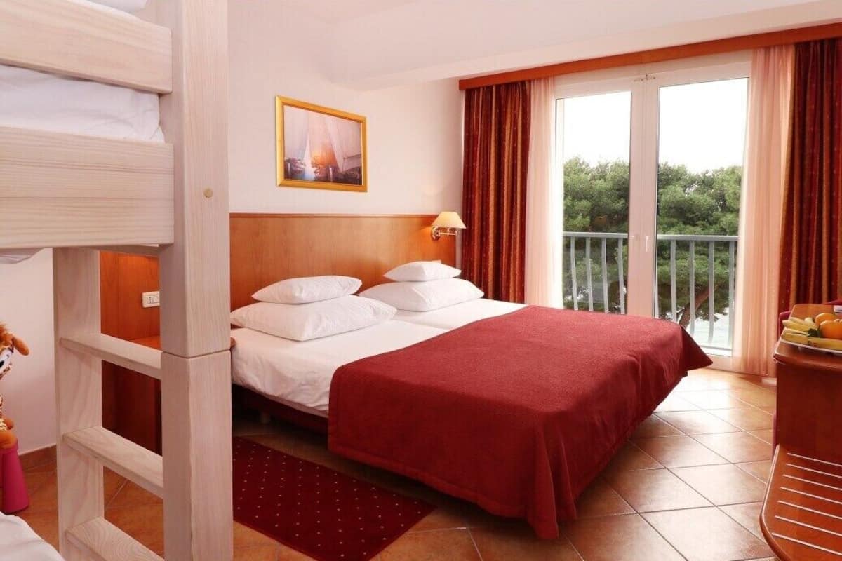 A hotel room with a bed, a wooden headboard, a red bedspread, a window with curtains, and a balcony with a railing.