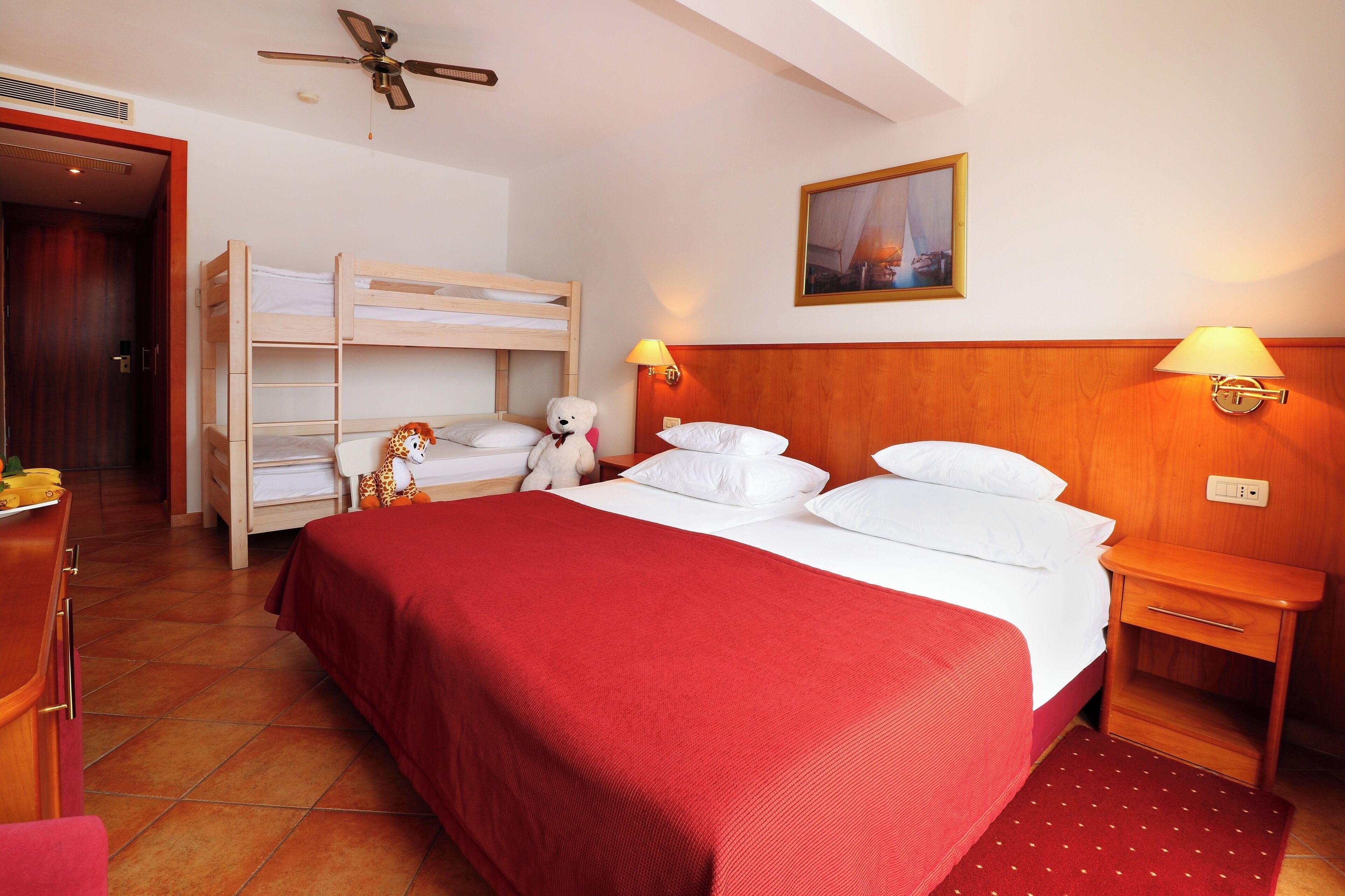 A hotel room with a bunk bed, a single bed with a red bedspread, a wooden nightstand with a lamp, a ceiling fan, and a painting on the wall.