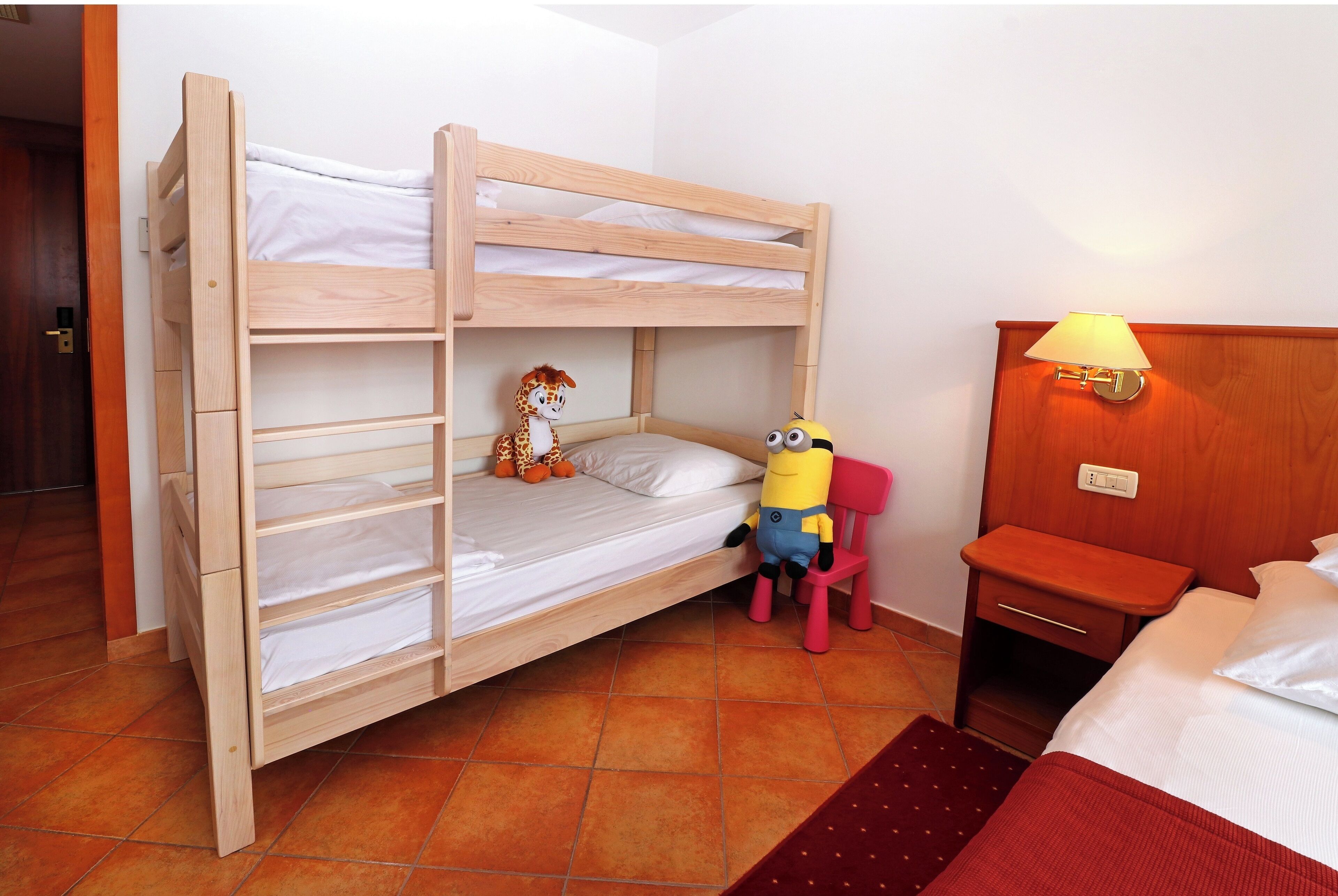 En køjeseng med en legetøjsgiraf og en Minion i underkøjen, et trænatbord med en lampe, og en rød stol.