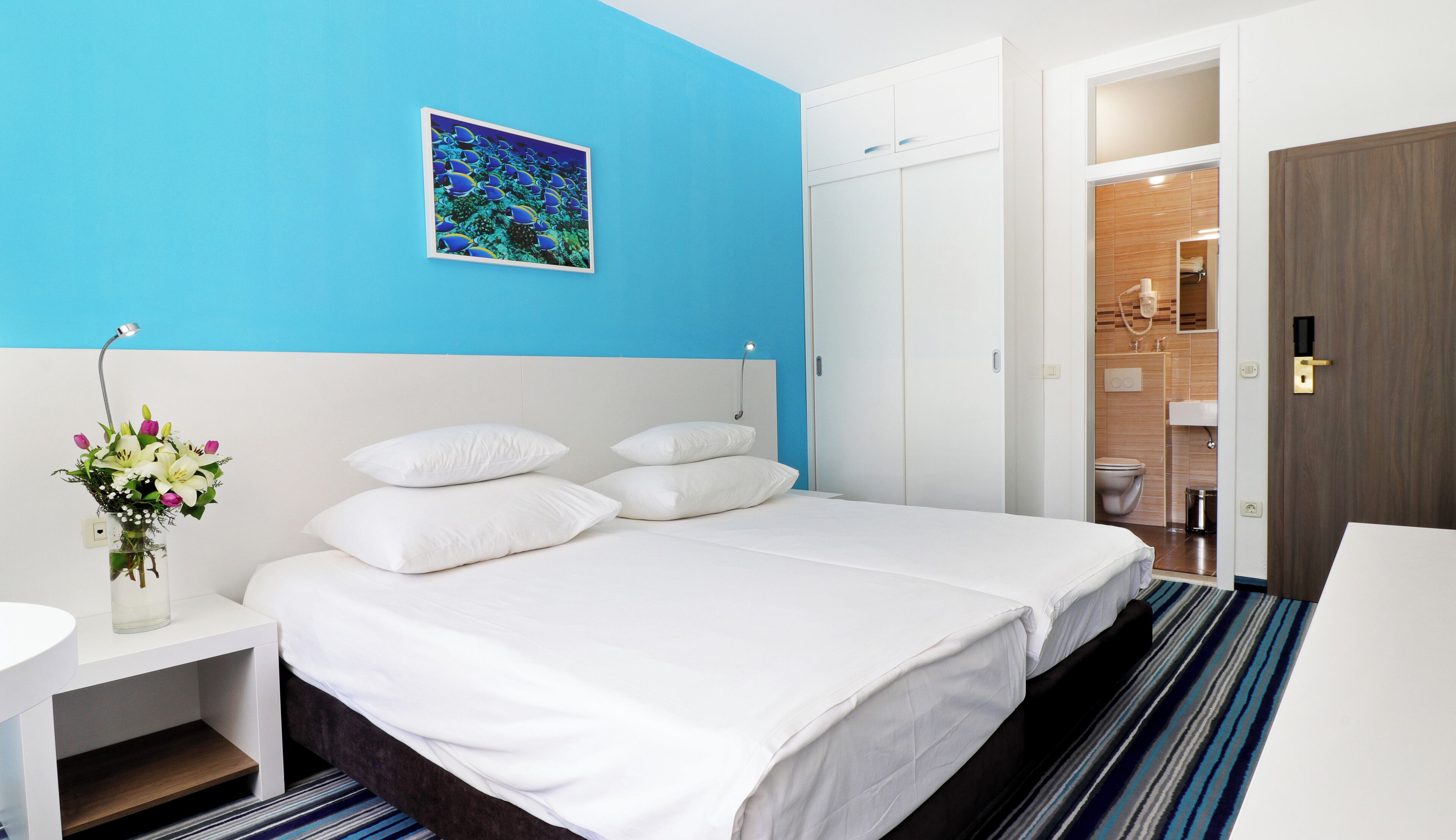 A hotel room with a large bed, white bedding, a bedside table with a vase of flowers, a blue accent wall, and a bathroom visible through an open door.