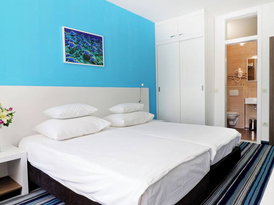 A hotel room with a large bed, white bedding, a bedside table with a vase of flowers, a blue accent wall, and a bathroom visible through an open door.