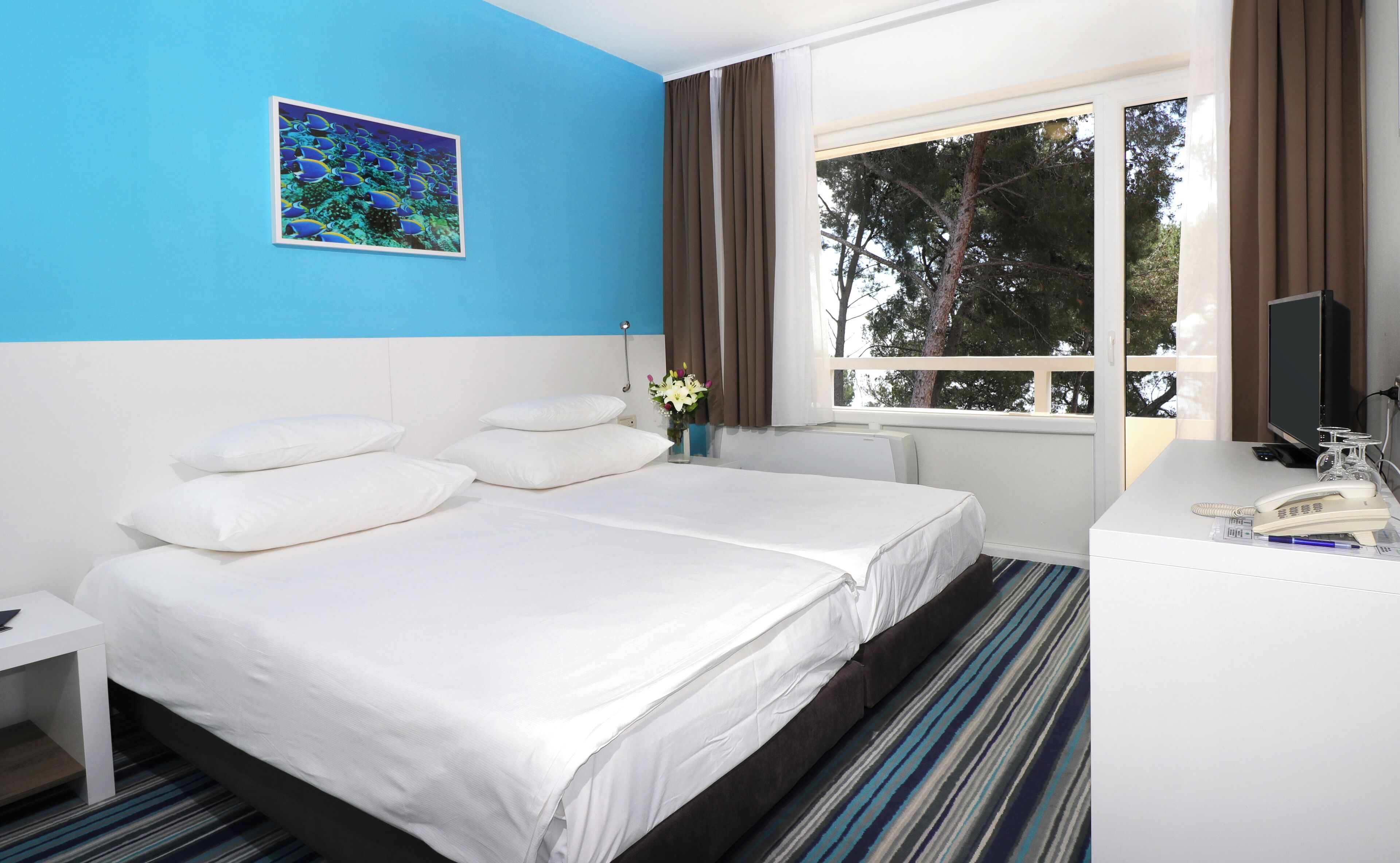 A hotel room with a bed, white pillows, a nightstand, a desk with a telephone, a computer monitor, and a window with curtains.