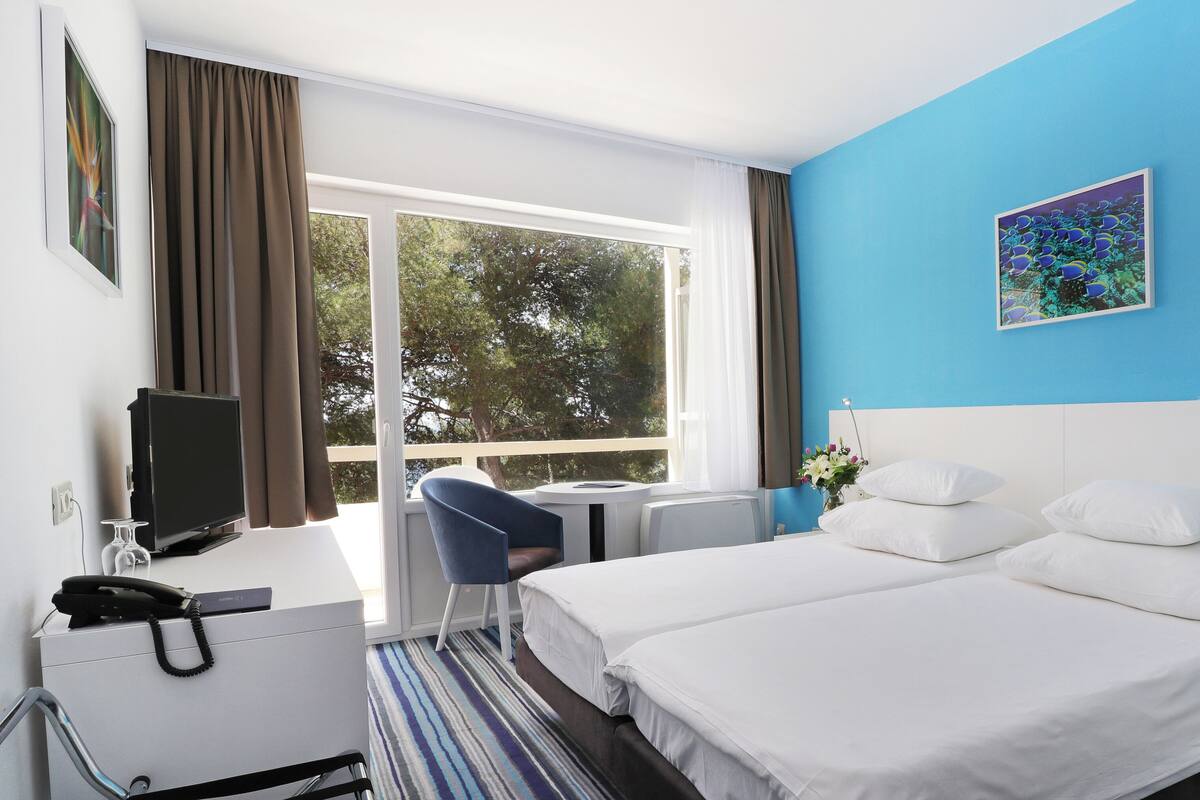 A hotel room with a large bed, a desk with a computer, a chair, and a window with curtains.