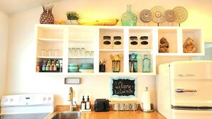 A kitchen with white cabinets, a white stove, and a white refrigerator. There are various kitchen items on the shelves, including jars, bottles, and a sign that says "Welcome to La Casita Bay". - Lake View Cottage (Three Mile Bay)