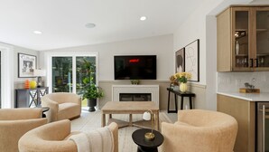 A modern living room with a fireplace, comfortable seating, a flat-screen TV, and a kitchen area with cabinets.
