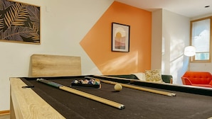 A pool table with balls and cues, a wooden headboard, a framed artwork, and a bright orange accent wall.