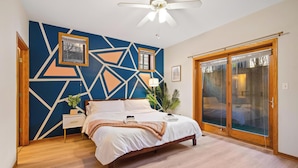 A bedroom with a geometric-patterned wall, a bed with white linens, a wooden floor, a ceiling fan, and a sliding glass door.
