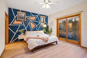 Une chambre avec un mur orné d’un motif géométrique, un lit recouvert de draps blancs, un plancher en bois, un ventilateur de plafond et une porte-fenêtre coulissante.