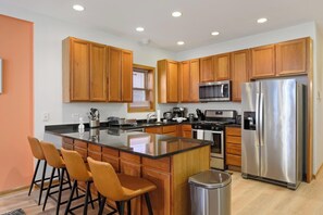 A modern kitchen with wooden cabinets, stainless steel appliances, a central island with bar stools, and a trash can.