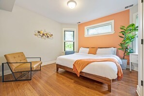 A bedroom with a wooden floor, a bed with white bedding, a brown chair, a potted plant, and a wall-mounted light fixture.