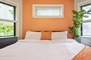 A bedroom with a bed, a window with blinds, a nightstand, and a potted plant.