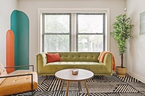 A room with a green sofa, a round table, a potted plant, and a window with blinds.
