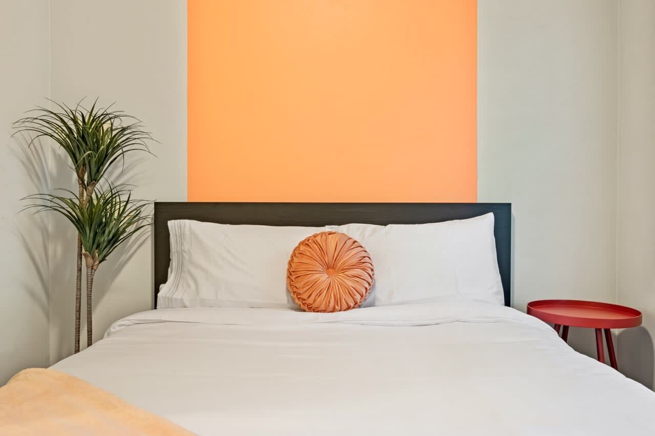 A bed with white bedding, a single orange decorative pillow, and a small red table beside it.