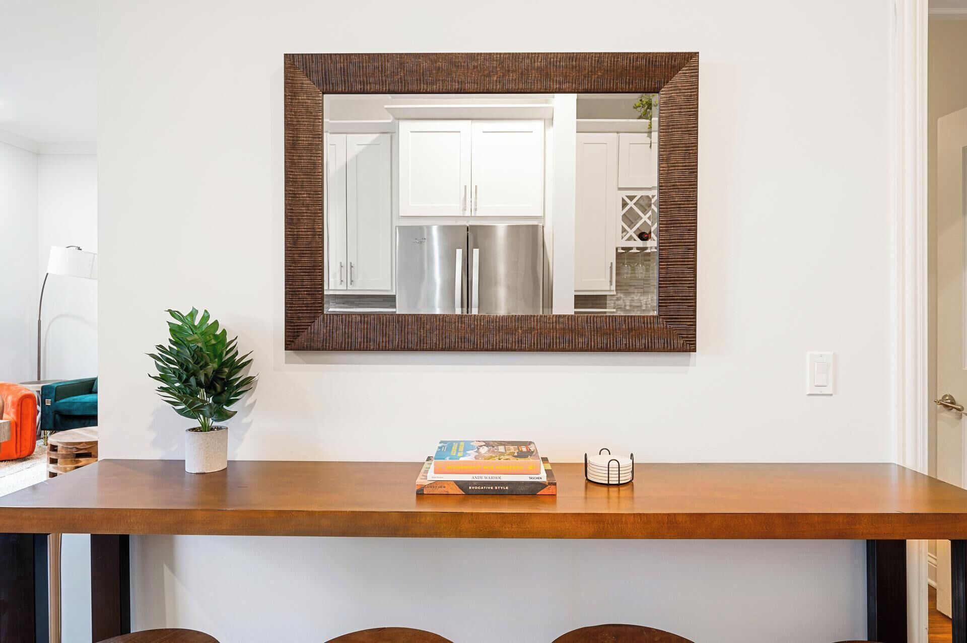 A modern kitchen with white cabinets and stainless steel appliances, a wooden dining table with a plant and a book, and a large mirror with a textured frame.