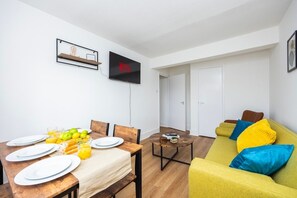 Uma sala de estar moderna com área de jantar, televisão de tela plana na parede e um sofá amarelo com almofadas coloridas.