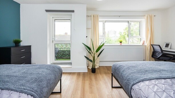 A bedroom with two beds, a desk, a chair, a radiator, and a window with curtains.