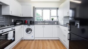 A modern kitchen with white cabinets, a black refrigerator, a washing machine, and a window with a view of the city.