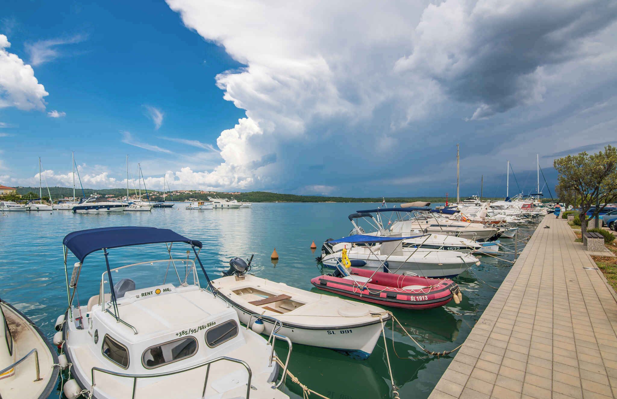 Un puerto deportivo con varias embarcaciones amarradas, un paseo marítimo pavimentado y una vista distante de la costa.
