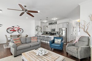 A modern living room with a gray sofa, blue armchair, and a wooden coffee table. A red bicycle is mounted on the wall. The kitchen area is visible in the background with white cabinets and a stainless steel refrigerator. - 🌊 Sundaze 30A – Walk to Beach-4 Bikes Included 🚲 (Santa Rosa Beach)