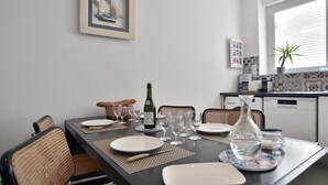 Un coin repas avec une table mise pour quatre personnes, comprenant des assiettes, des verres et une bouteille de vin. On y trouve des chaises en osier et un tableau encadré accroché au mur.