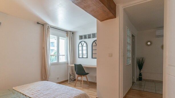 Un dormitorio con cama, escritorio y silla. Hay una ventana con cortinas, una puerta que da a otra habitación y una planta.