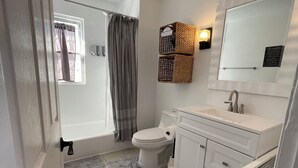 Une salle de bain comprenant des toilettes, un lavabo et une baignoire. On y trouve une fenêtre avec des rideaux, un porte-serviettes et un miroir.