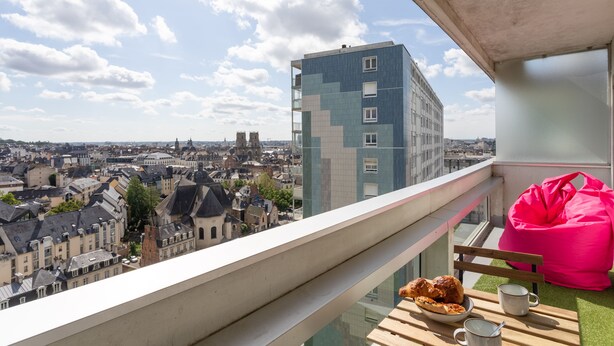 A balcony with a view of a cityscape, a pink bean bag, a wooden table with pastries and a mug, and a tiled building in the background. - Le Studio de l'Ange - Vue imprenable (Rennes)