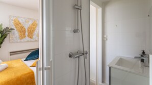 A modern bathroom with a glass shower enclosure, a white sink, and a bed with a yellow blanket and blue pillow. - Le Studio de l'Ange - Vue imprenable (Rennes)