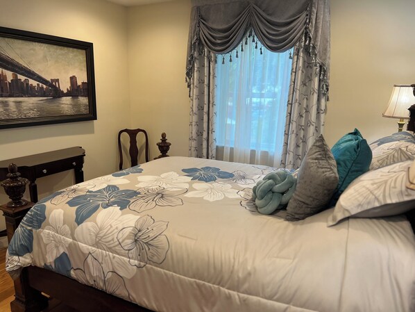 Un dormitorio con una cama grande, ropa de cama floral, un cuadro de paisaje urbano, una ventana con cortinas y una mesita de noche con lámpara.